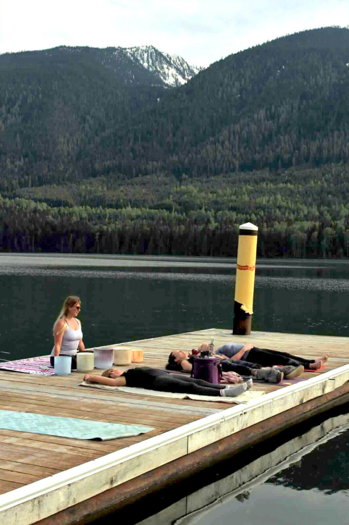 Sandy sits and leads yoga on the dock as several women lay on mats in front of her. Below them is the lake and in the background are tree-covered mountains.