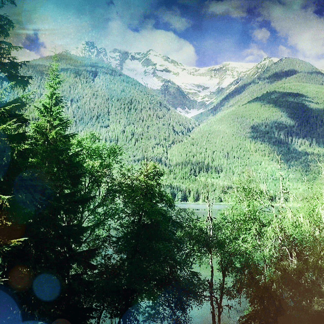 Three mountains covered in leafy trees with a lake at the base and a handful of fluffy clouds in the bright blue sky.