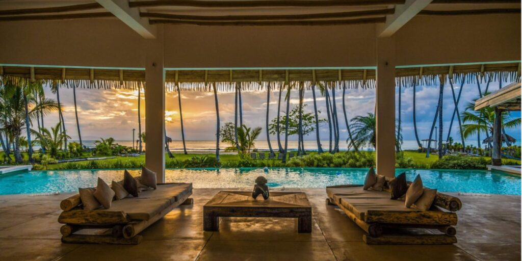 Wicker table and chairs in front of a pool with palm trees and a sunset in the background.