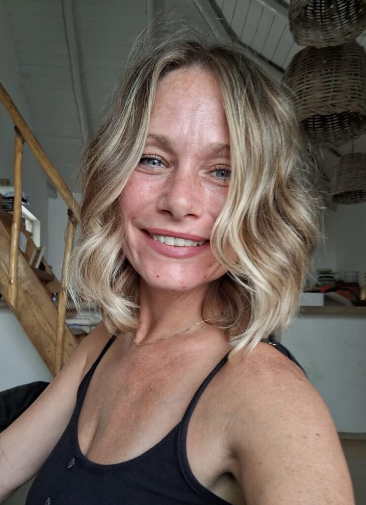 Rebecca is a woman with a light skin tone and wavy blonde shoulder-length hair. She smiles warmly and wears a black tank top.