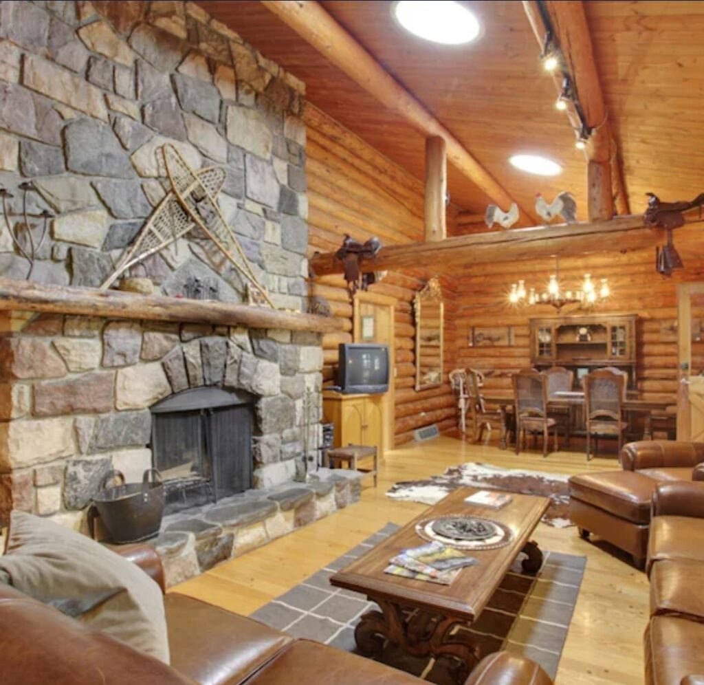 The inside of a log cabin with leather couches, a wooden dining table, and a large stone fire place.