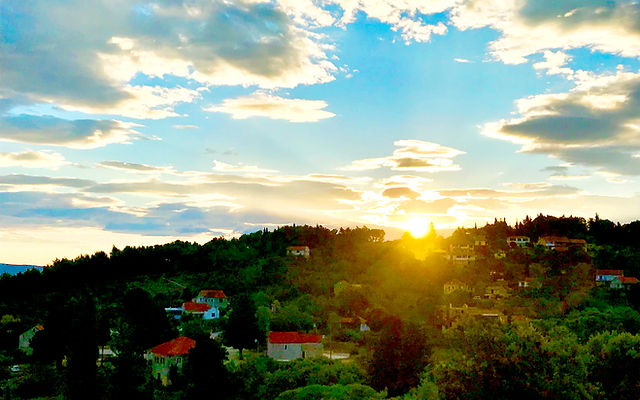A tree-covered hillside dappled with red-roofed houses. In the background a sun sets in a partially cloudy blue sky.