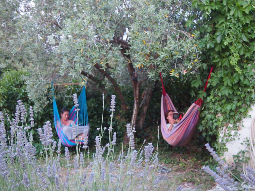 Two women sit and read in colorful hammocks. Around them are flowers and green-leafed trees.