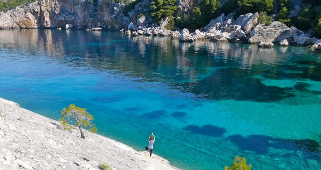 A person does yoga at the water's edge of a low-sloping granite hill. The wide stream of water is bright teal and aqua, and on the opposite bank are low rocky cliffs.