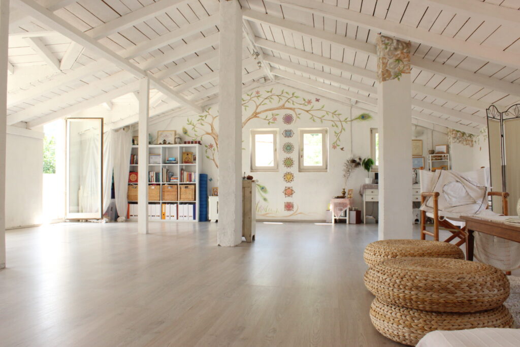 A large room with white walls and exposed rafters. On the wall is a chakra and tree mural, and there is natural-materials furniture against the walls.