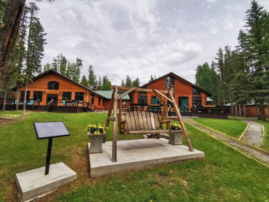 A two-person swinging wooden chair sits on a green-grass lawn in front of a large red-painted cabin.