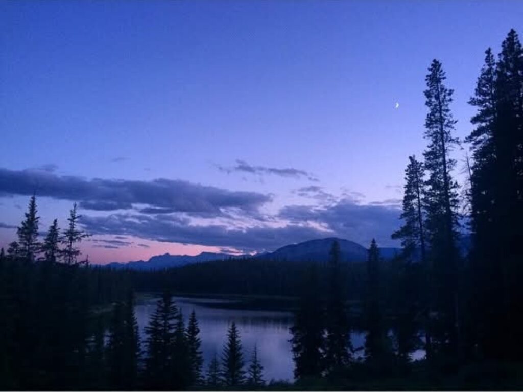 Sun sets over a low mountain. In the foreground is a tree-lined lake.