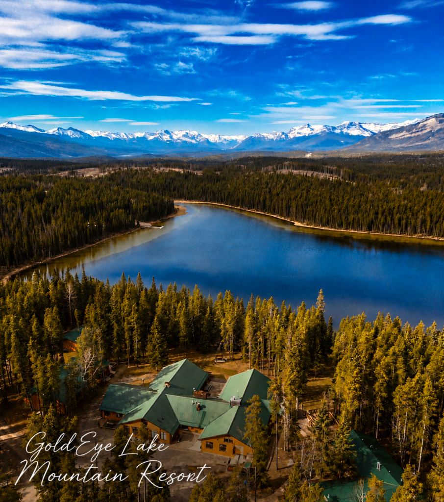 A calm blue lake amid pine tree forests. In the image foreground, a green-roofed lodge sits among the trees. The sky is blue with wisps of white clouds and snow-capped mountains stand tall in the background.