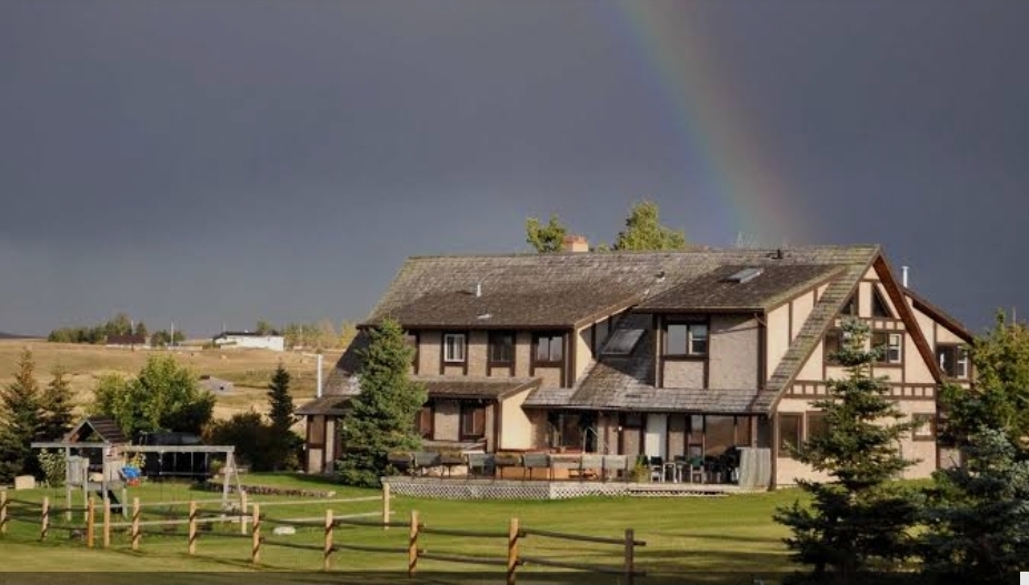 Large timber-framed lodge-style house with a spacious lawn, fenced yard, evergreen trees, and a rainbow in the dark sky.